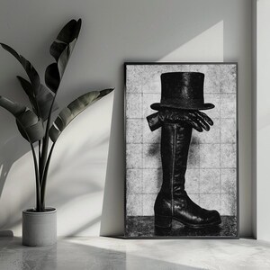 Op de afbeelding: Zwart-wit ingelijste kunstdruk met een hoge hoed, handschoenen en een hoge laars. Het kunstwerk wordt gepresenteerd tegen een achtergrond met textuur, met een plant in een pot op de voorgrond.