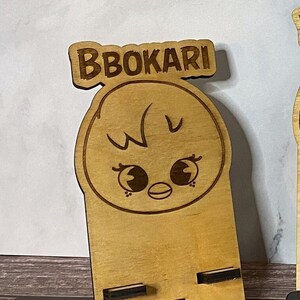 May include: A wooden phone stand with a cartoon chick face and the name "BBOKARI" engraved on top. The stand has two slots for holding a phone.