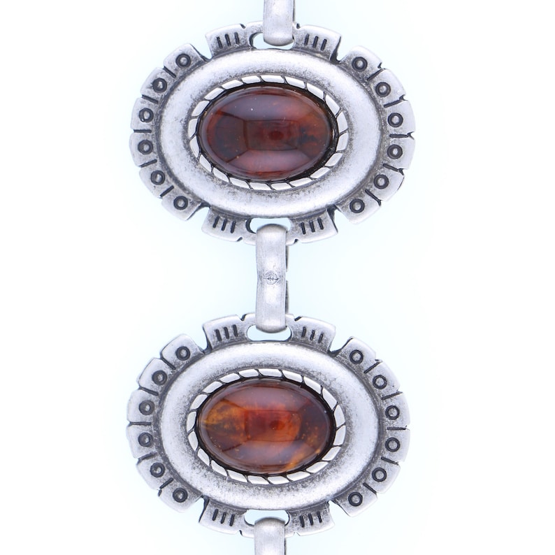 May include: Close-up of a silver-toned bracelet featuring two oval-shaped, amber-colored stones. Each stone is set within a decorative frame with a textured border and a series of small, circular accents. The bracelet links are connected by small, curved bars.