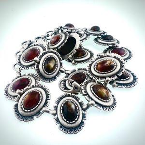 May include: A silver-toned belt with oval-shaped, reddish-brown and brown stones. The belt features a series of decorative, floral-inspired links and a clasp closure. The stones are set within a detailed, textured metal frame.