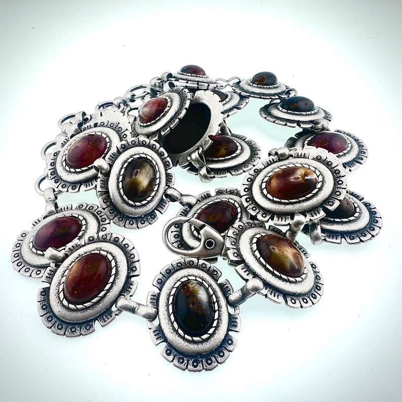 May include: A silver-toned belt with oval-shaped, reddish-brown and brown stones. The belt features a series of decorative, floral-inspired links and a clasp closure. The stones are set within a detailed, textured metal frame.