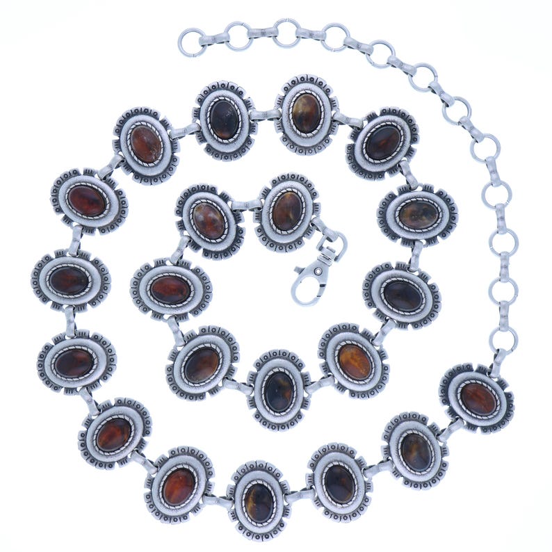 May include: Silver-toned metal belt with oval, brown-toned stones set in decorative frames. The belt features a chain extension and a clasp closure. The design is intricate and detailed, suitable for adding a touch of flair to any outfit.