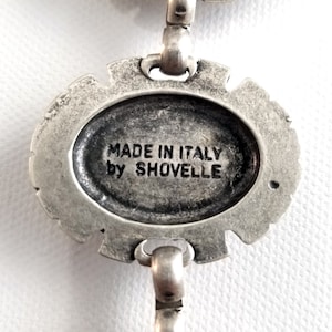 May include: Close-up of a silver-toned metal pendant with an oval center. The text "MADE IN ITALY by SHOVELLE" is inscribed within the oval. The pendant has a decorative, ornate border and is connected to a chain.