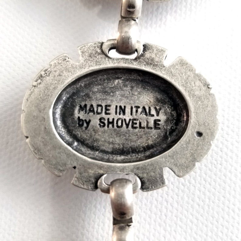 May include: Close-up of a silver-toned metal pendant with an oval center. The text "MADE IN ITALY by SHOVELLE" is inscribed within the oval. The pendant has a decorative, ornate border and is connected to a chain.