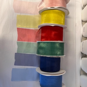 May include: A collection of eight spools of sheer ribbon in various colours, including pink, yellow, red, green, blue, and light blue. The ribbon is wrapped around white cardboard spools.