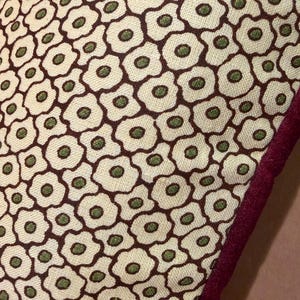May include: A textile fabric featuring a repeating pattern of stylized off-white flowers with dark brown outlines and small green centers. The fabric has a visible woven texture and is finished with a thick, dark maroon velvet-like edge trim.