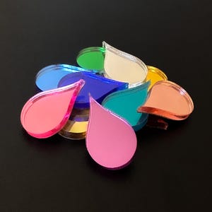 May include: A selection of multi-coloured, teardrop-shaped acrylic pieces. The pieces are in shades of pink, blue, green, and gold. They have a mirrored surface, creating a vibrant display. The shapes are scattered on a dark surface.