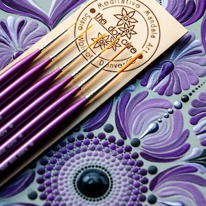 May include: A collection of purple dotting tools with silver tips, displayed on a wooden plaque with the text "Meditative Mandala Art" and floral motifs. The background showcases a detailed mandala design in shades of purple, grey, and white.