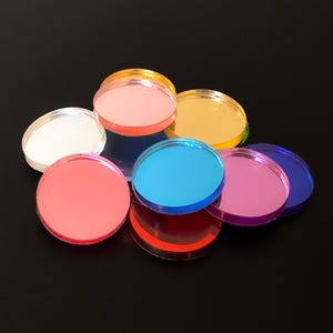 May include: A selection of multi-coloured, circular acrylic discs in shades of pink, blue, yellow, and purple. The discs are stacked and scattered, creating a vibrant display against a dark background. The transparent nature of the discs allows light to pass through, enhancing their colours.