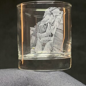 Peut inclure: Un verre &agrave; whisky transparent avec une image grav&eacute;e d'une personne en combinaison de vol. L'image est d&eacute;taill&eacute;e et semble &ecirc;tre un portrait. Le verre repose sur une surface gris fonc&eacute;.