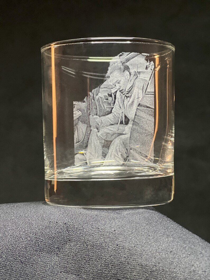 Peut inclure: Un verre &agrave; whisky transparent avec une image grav&eacute;e d'une personne en combinaison de vol. L'image est d&eacute;taill&eacute;e et semble &ecirc;tre un portrait. Le verre repose sur une surface gris fonc&eacute;.