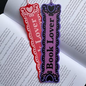 May include: Two decorative bookmarks. One is red with the word "Lover" and a heart design. The other is purple with the words "Book Lover" and a heart design. Both have a pink string.
