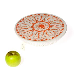 May include: A white bowl cover featuring an orange sunflower pattern. A green apple sits beside the cover. The cover has a tag that says "halo".