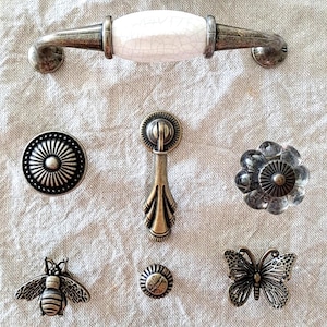 May include: Assortment of antique-style cabinet hardware in various shapes and sizes, including a butterfly, bee, flower, and round knobs, a pull handle with a white ceramic insert, and a cup pull.