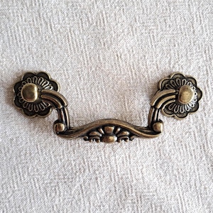 May include: Aged brass drawer pull with a floral design. The pull features a curved handle and decorative floral accents. The hardware has a warm, aged brass colour, suitable for vintage or rustic furniture.