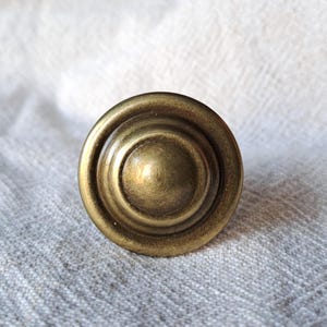 May include: A close-up of a round, antique brass cabinet knob. The knob has a tiered design with concentric circles and a slightly domed centre. The finish has a warm, aged appearance.