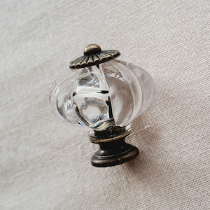 May include: A clear glass cabinet knob with a pumpkin-shaped, ribbed design. The knob features an antique brass-toned metal base and a matching circular top cap with radial grooves, providing a decorative hardware element for furniture.