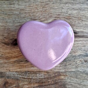 May include: A heart-shaped ornament in a pale lavender colour, set against a wooden background. The heart has a glossy finish, reflecting light. The wooden surface displays a natural grain pattern. The object is likely a decorative piece.
