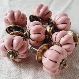 May include: A selection of pink ceramic cabinet knobs, each with a pumpkin shape and gold detailing. The knobs have brass-toned bases and are suitable for furniture and home decoration. Each knob is approximately 4 cm in diameter.