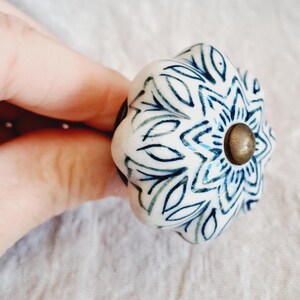May include: A white ceramic cabinet knob with a floral design in blue and green. The knob has a brass-coloured centre and a rounded shape. The floral pattern is symmetrical and detailed, adding a decorative touch to furniture.