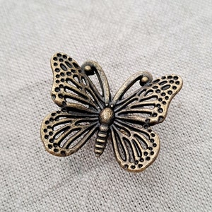 May include: A gold-toned metal butterfly charm with a delicate, intricate design. The butterfly has a detailed body and wings with a cutout pattern.