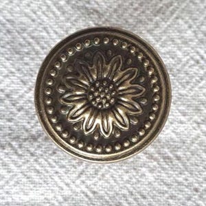 May include: A round metal cabinet knob with an antique brass finish. The face of the knob features a raised, embossed floral design in the centre, surrounded by a circular border of small, evenly spaced raised dots along the outer edge.