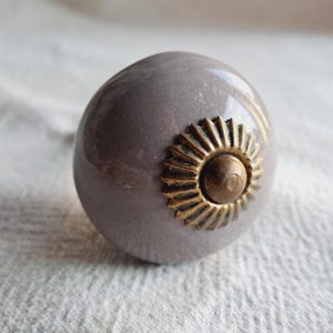 May include: A grey ceramic cabinet knob with a brass sunflower design. The knob has a smooth, rounded shape and a glossy finish. The brass detail adds a touch of elegance to the hardware.
