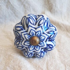 May include: A ceramic cabinet knob with a floral pattern in white and blue. The knob features a brass-coloured centre and a rounded, flower-like shape. The background is a light-coloured, textured fabric.