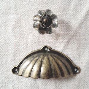 May include: A clear, faceted glass knob with a circular bronze-toned metal centre and a matching bronze-toned metal cup pull handle featuring a scalloped, shell-like design with three screw holes for mounting hardware.