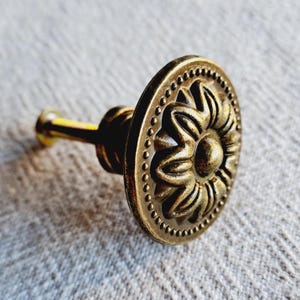 May include: A close-up of a bronze cabinet knob. The knob has a detailed floral design in the centre, surrounded by a dotted border. The hardware has a vintage aesthetic, with a long screw for installation.