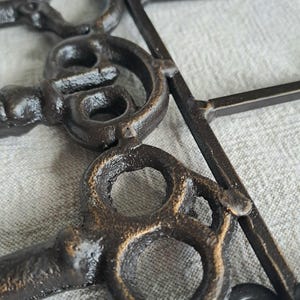 May include: Close-up of a decorative metal key-shaped object. The keys are a dark bronze colour with a textured finish, and are connected to a dark metal frame. The background is a light grey fabric.