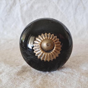 May include: A round, glossy black cabinet knob featuring a central brass-toned metal accent. The metal piece has a scalloped, sunburst-like edge surrounding a smooth, raised circular centre, providing a contrast to the dark, reflective surface.