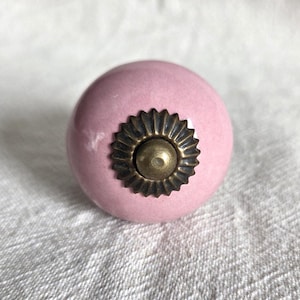 May include: A round, smooth, light pink ceramic cabinet knob featuring a central antique brass-toned metal accent piece. The metal component has a scalloped, sunburst-like edge surrounding a small, raised, circular dome in the centre.