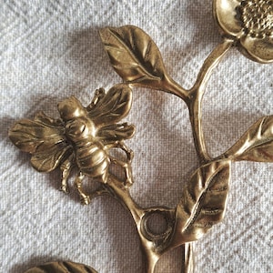 May include: A golden metal decorative item with a bee and flower design. The bee is positioned on a branch with leaves and a flower. The piece has a vintage appearance and is made of metal.