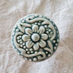 May include: A round, ceramic cabinet knob with a floral design. The knob is a pale teal colour with raised white detailing. The design includes a central flower, leaves, and small beaded accents. The knob is likely used for furniture or cabinetry.