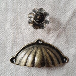 May include: A clear, flower-shaped glass cabinet knob with a bronze base sits above a bronze, shell-shaped drawer pull. Both pieces of hardware are set against a neutral, textured background.