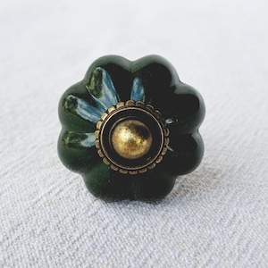 May include: A dark green ceramic cabinet knob with a flower-like design. The knob features a brass-coloured centre and a decorative brass ring. The knob is approximately 5 cm in diameter.