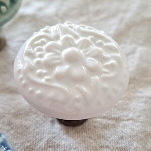 May include: White ceramic cabinet knob with a raised floral design. The knob has a white finish and a slightly pink hue.