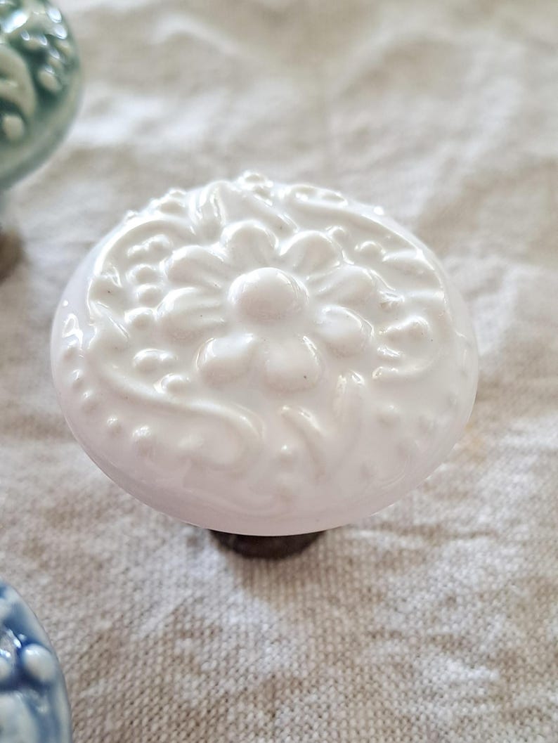 May include: White ceramic cabinet knob with a raised floral design. The knob has a white finish and a slightly pink hue.