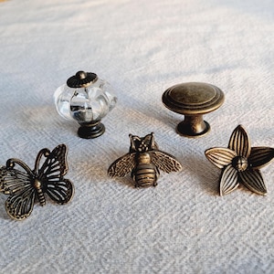 May include: A selection of decorative cabinet knobs. The knobs feature a clear glass design, a round bronze knob, a butterfly, a bee, and a flower. All have a bronze finish and are displayed on a neutral background.