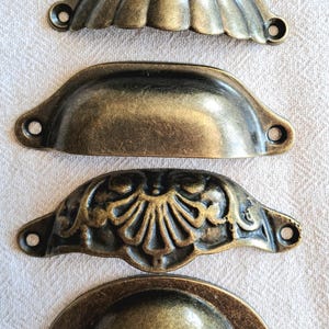 May include: Four antique bronze cabinet pulls in a half-moon shape. The pulls have different designs, including fluted and floral patterns, and a smooth curved surface. Each pull has two holes for mounting. The pulls are in a bronze colour.