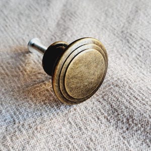 May include: A bronze cabinet knob with a circular design. The knob has a textured surface and a silver stem. The hardware is designed for use on drawers or cabinet doors.