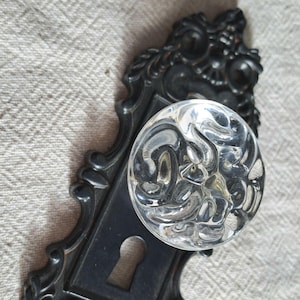 May include: An antique door knob with a clear, textured glass knob and a black, ornate metal backplate. The backplate features a keyhole and decorative scrollwork. The knob is round and reflects light, showcasing its intricate design.