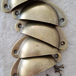 May include: Five antique brass cup pulls with a set of screws. The cup pulls are arranged in a stack, with the largest at the bottom and the smallest at the top.