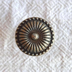 May include: Close-up of a bronze cabinet knob with a circular, sunburst design. The knob has a central raised circle, radiating lines, and a beaded outer edge. The hardware is likely for furniture.