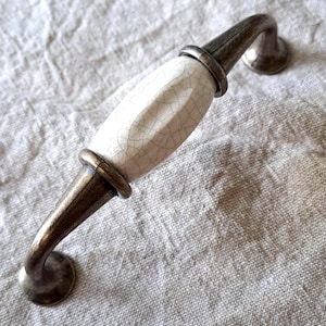 May include: A cabinet handle featuring a white, crackled ceramic centre and bronze-coloured metal hardware. The handle has a curved design. The hardware is suitable for drawers and cabinets. The handle is set against a neutral linen background.