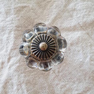 May include: Clear glass cabinet knob with a flower-like design. The knob features a central, circular, antique-bronze-coloured base with radiating lines, surrounded by clear, rounded glass petals. The knob is designed for furniture and home decor.