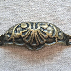 May include: An antique brass-effect drawer pull with an ornate, symmetrical design. The pull has a curved shape with decorative floral and scrollwork details. Two holes are on each end for mounting. The finish has a vintage, aged appearance.