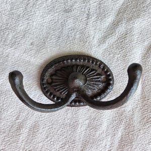 May include: A dark brown, cast iron double wall hook featuring an oval backplate with a decorative sunburst pattern and a beaded border. The hook has two curved arms extending outward, each ending in a rounded tip, with two visible screw holes.