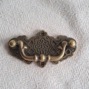 May include: Ornate antique brass drawer pull. The decorative backplate has a lattice pattern and a central floral design. The curved handle has ball-shaped ends. The pull is suitable for furniture.